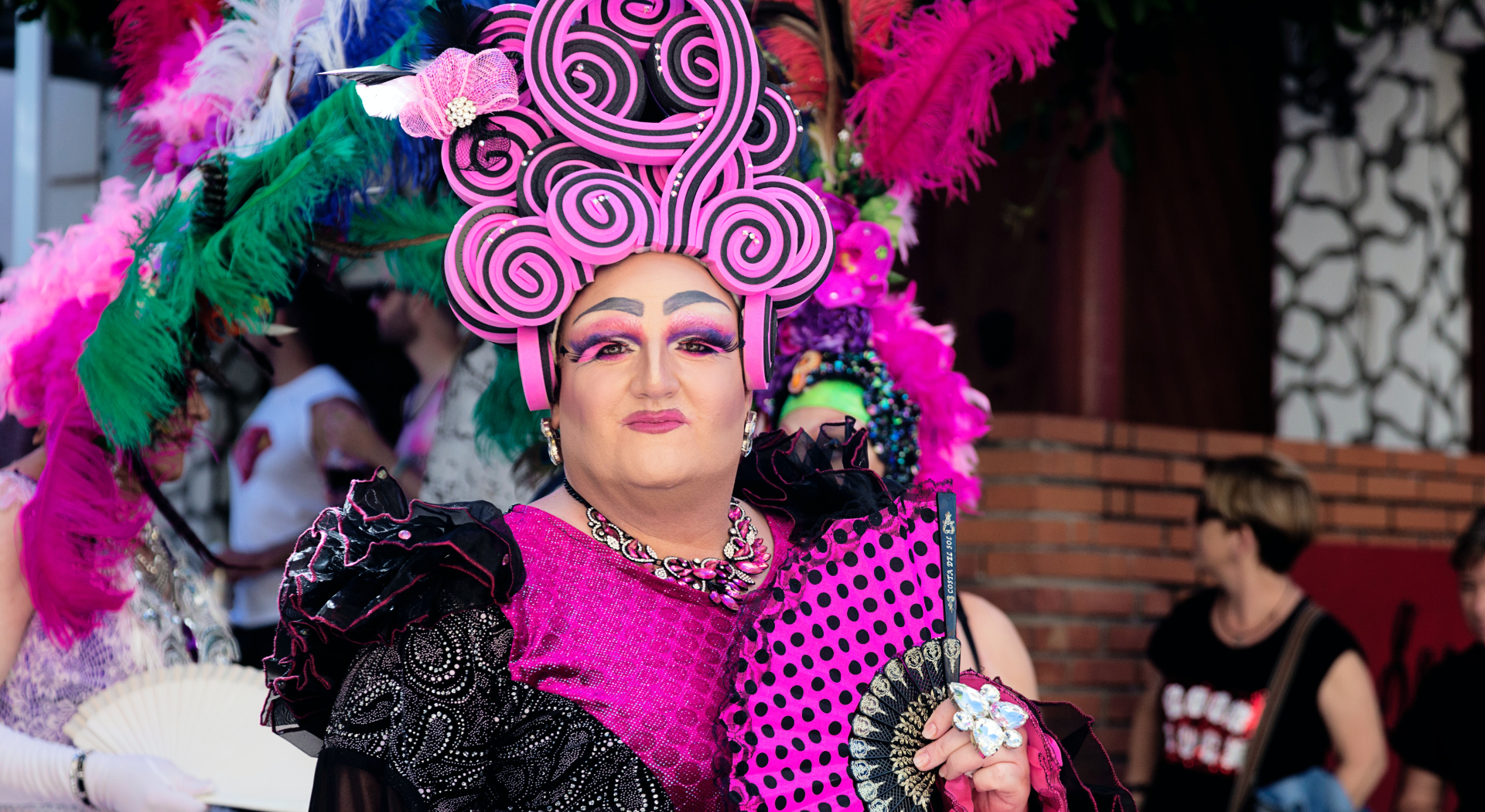 A drag queen dressed in vibrant colours of mostly pink with black polka dots. She is wearing pink eyeshadow and has a pink head piece on top of her head.