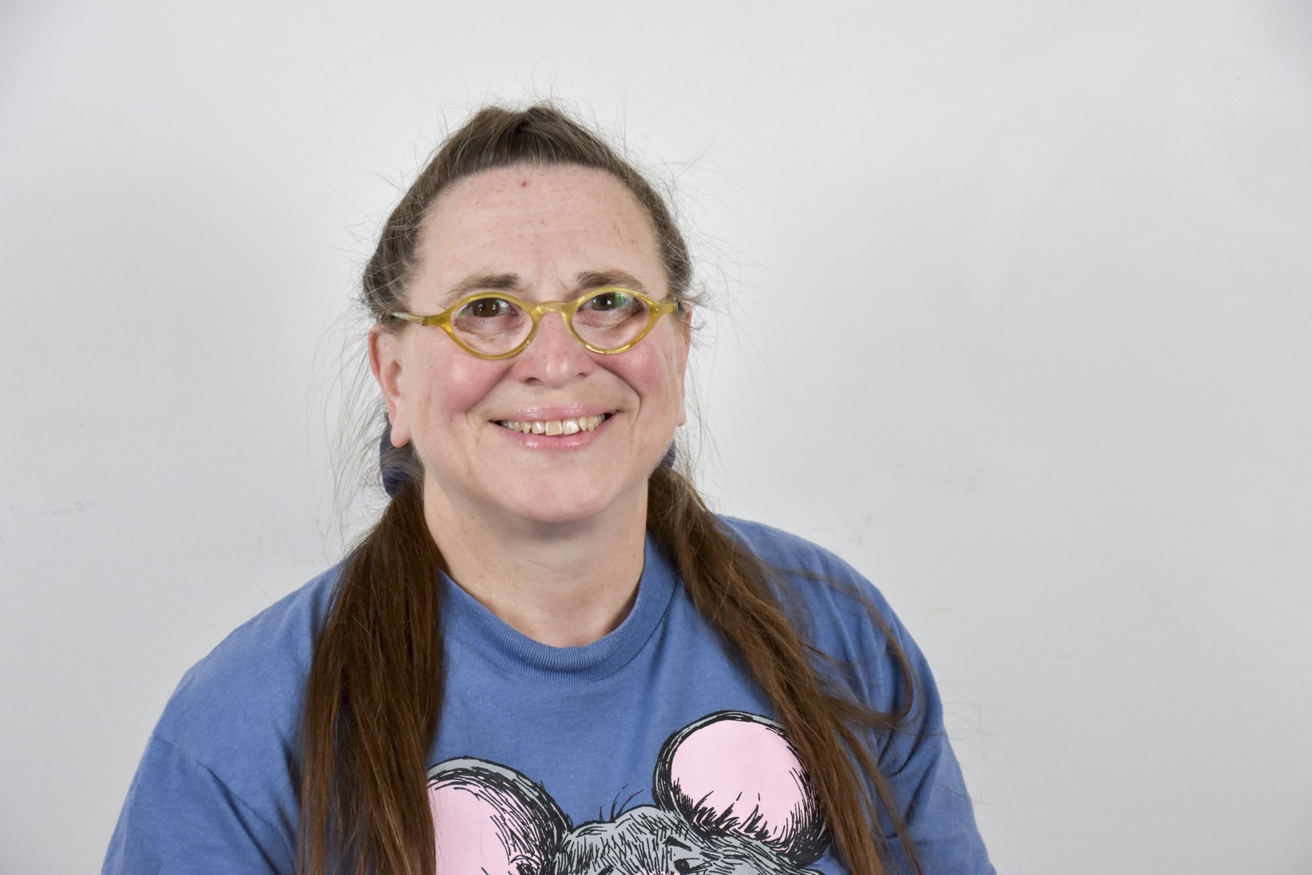 Photo of Kathy Austin. She is wearing a blue shirt, with her brown hair styled in a half up, half down hair style. She is wearing glasses and smiling at the gamer. The background is a light grey wall.
