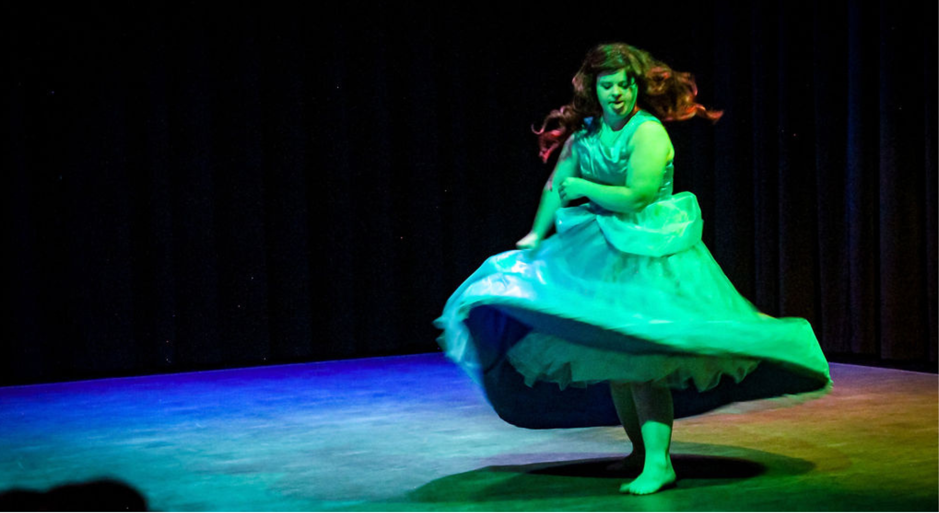 An image of an NaAC performer spinning in a costume on stage with blue light casted on the left side and a green light highlighting the right side of the stage and performer.